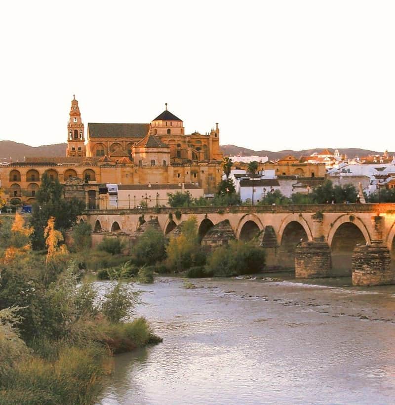 A historic stone bridge crosses a river, leading to a large, ornate building with arches and a tall tower in the background.