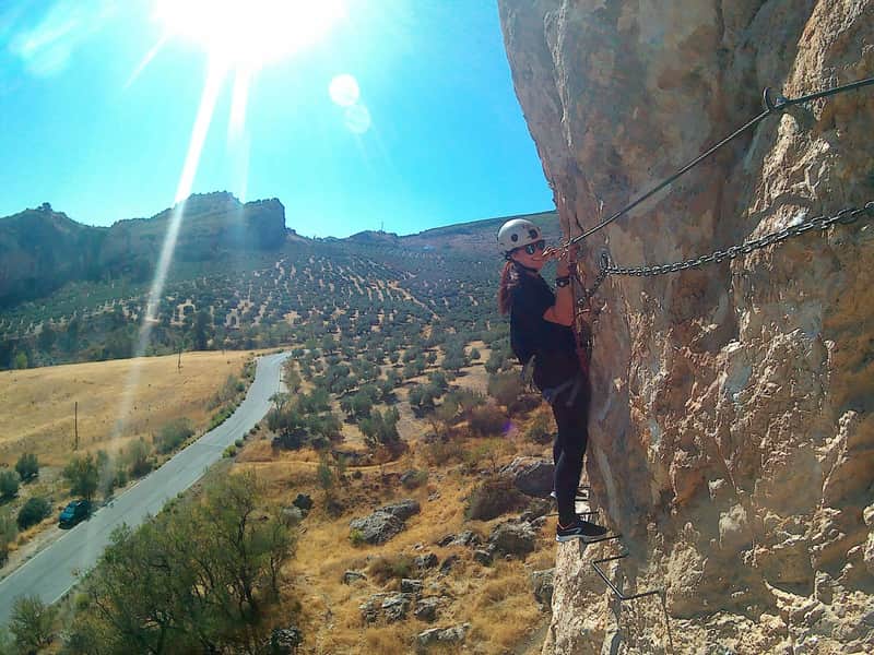A person in climbing gear ascends a rocky cliff, with a sunny landscape and a winding road in the background.