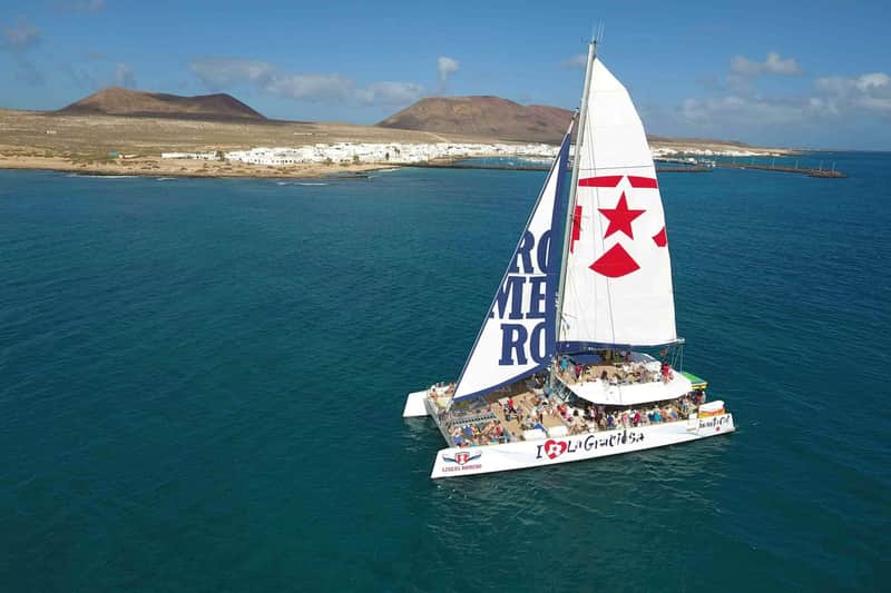 Catamaran with "I ❤️ La Graciosa" and star on sails, sailing in blue water near a coastal town with white buildings and hills.