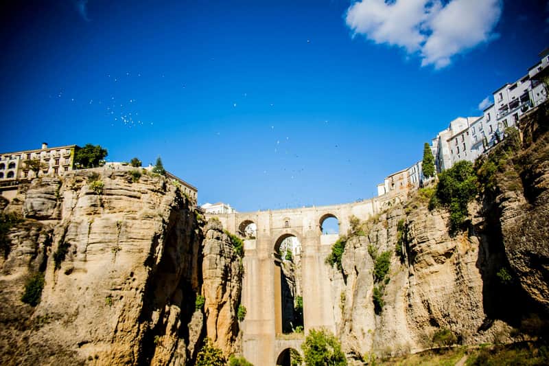 Ronda: Guided Tour from Málaga Tall stone bridge with multiple arches spanning a deep canyon, with birds flying in the blue sky and cliffs covered in greenery.