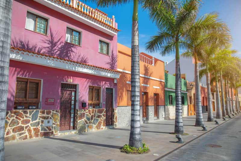 Colorful buildings with a pink and orange facade, palm trees lining the street, under a clear blue sky.
