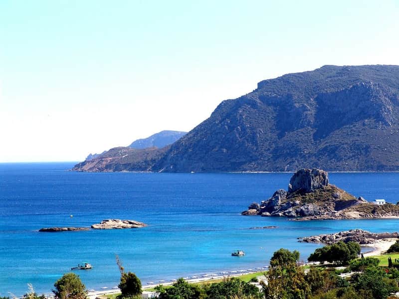 A coastal landscape with blue water, rocky hills in the background, small islands, and trees in the foreground.