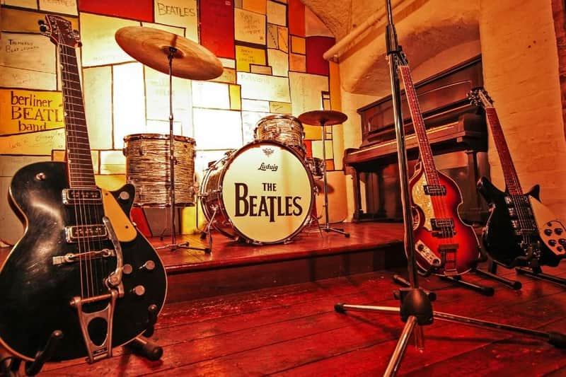 Una batteria vintage con il logo dei "Beatles", delle chitarre e un pianoforte su un palco di legno con pannelli colorati alle pareti.