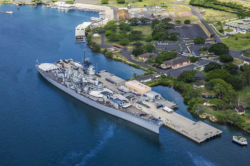 Aerial view of a large battleship docked at a pier surrounded by greenery and buildings near a waterfront.