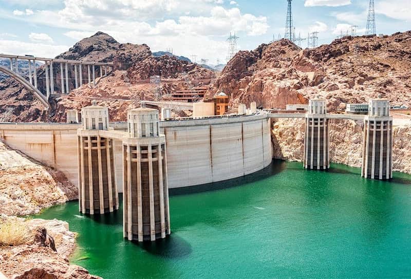 A large dam with towers spans a green reservoir, surrounded by rocky terrain, with electrical infrastructure and a blue sky.