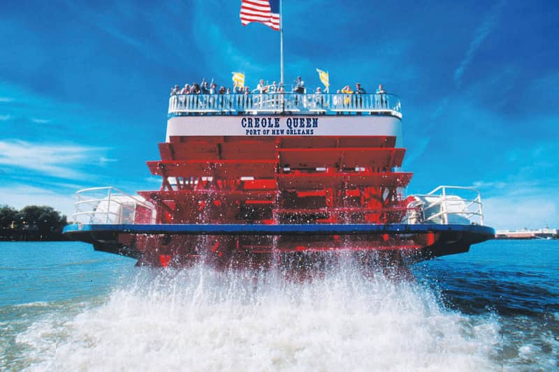 Red paddlewheel of the Creole Queen riverboat, with people on the upper deck and an American flag above against a clear blue sky.
