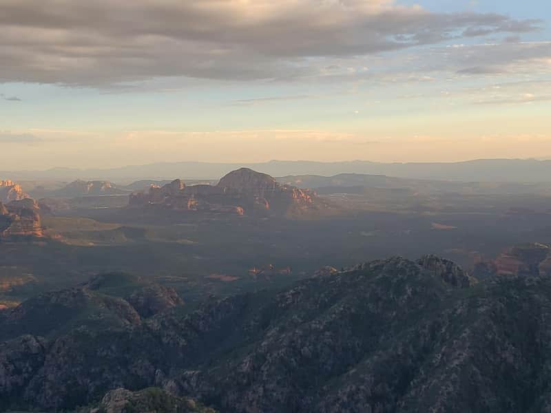 Mountainous landscape with rocky peaks, a distant mesa, and a sky filled with clouds during sunset.