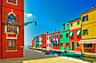 Case colorate costeggiano un canale con piccole barche attraccate, sotto un cielo blu brillante. Un palo della luce si estende da un edificio sulla sinistra.
